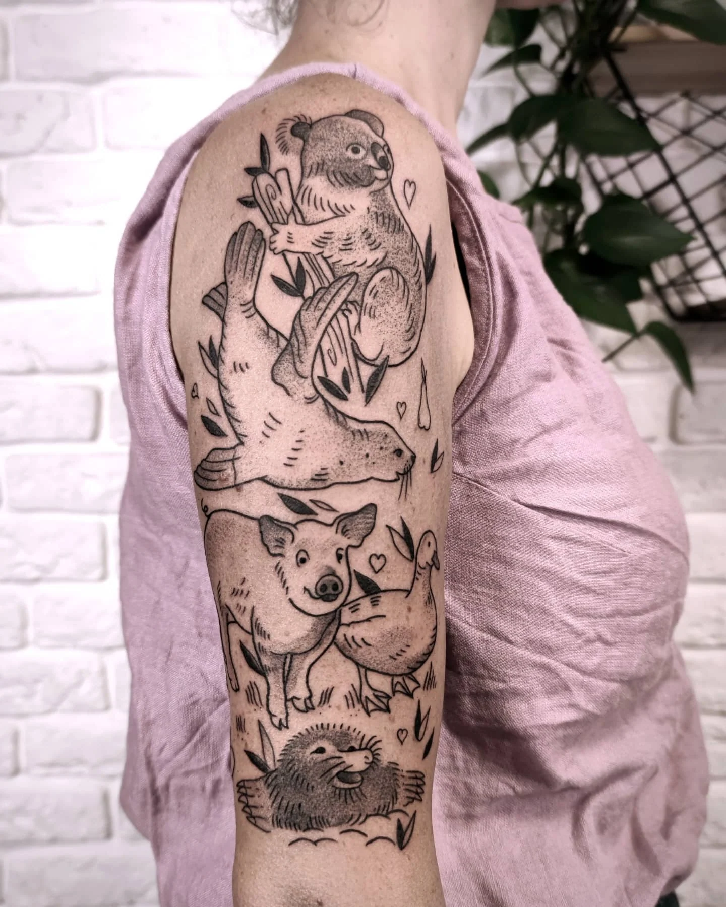 Zbliżenie na rękaw tatuażu przedstawiający kilka uroczych zwierząt, między innymi koalę, świnię i jeżozwierza, uzupełnionych liśćmi i drobnymi sercami.