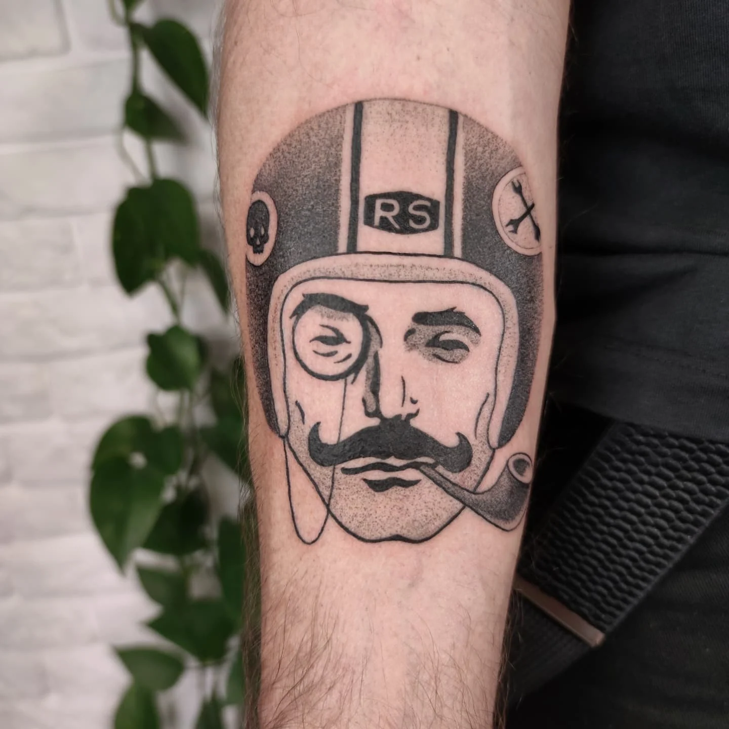 Tatuaż na przedramieniu przedstawiający stylizowaną twarz w kasku motocyklowym, z monogramem „RS” powyżej; obok mężczyzna prezentuje tatuaż siedząc na motocyklu., zdjęcie 1 z 2