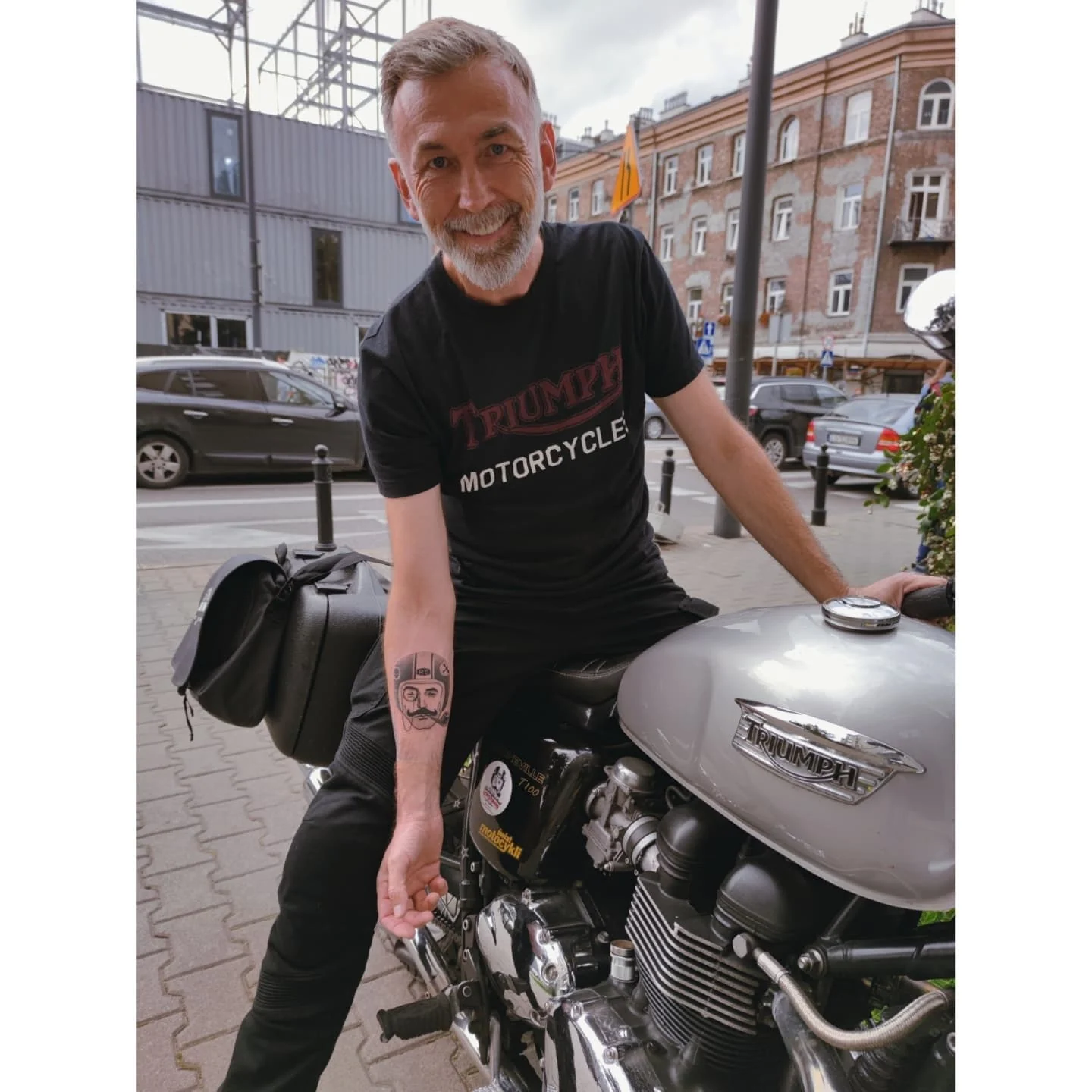 Tatuaż na przedramieniu przedstawiający stylizowaną twarz w kasku motocyklowym, z monogramem „RS” powyżej; obok mężczyzna prezentuje tatuaż siedząc na motocyklu., zdjęcie 2 z 2