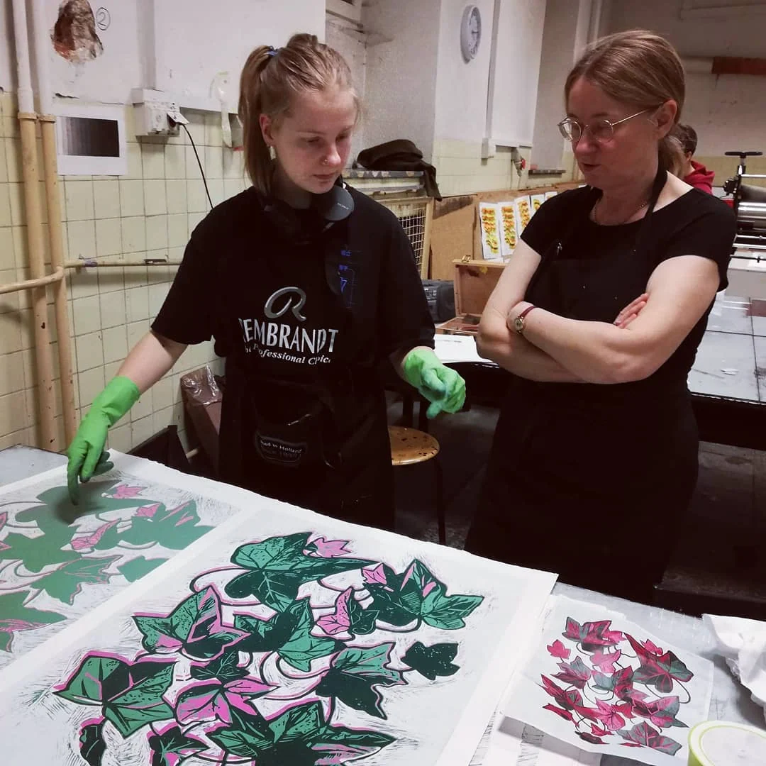Dwie osoby pracują przy botanicznym linorycie przedstawiającym liście i kwiaty; na stole leżą kolorowe odbitki oraz narzędzia do druku.