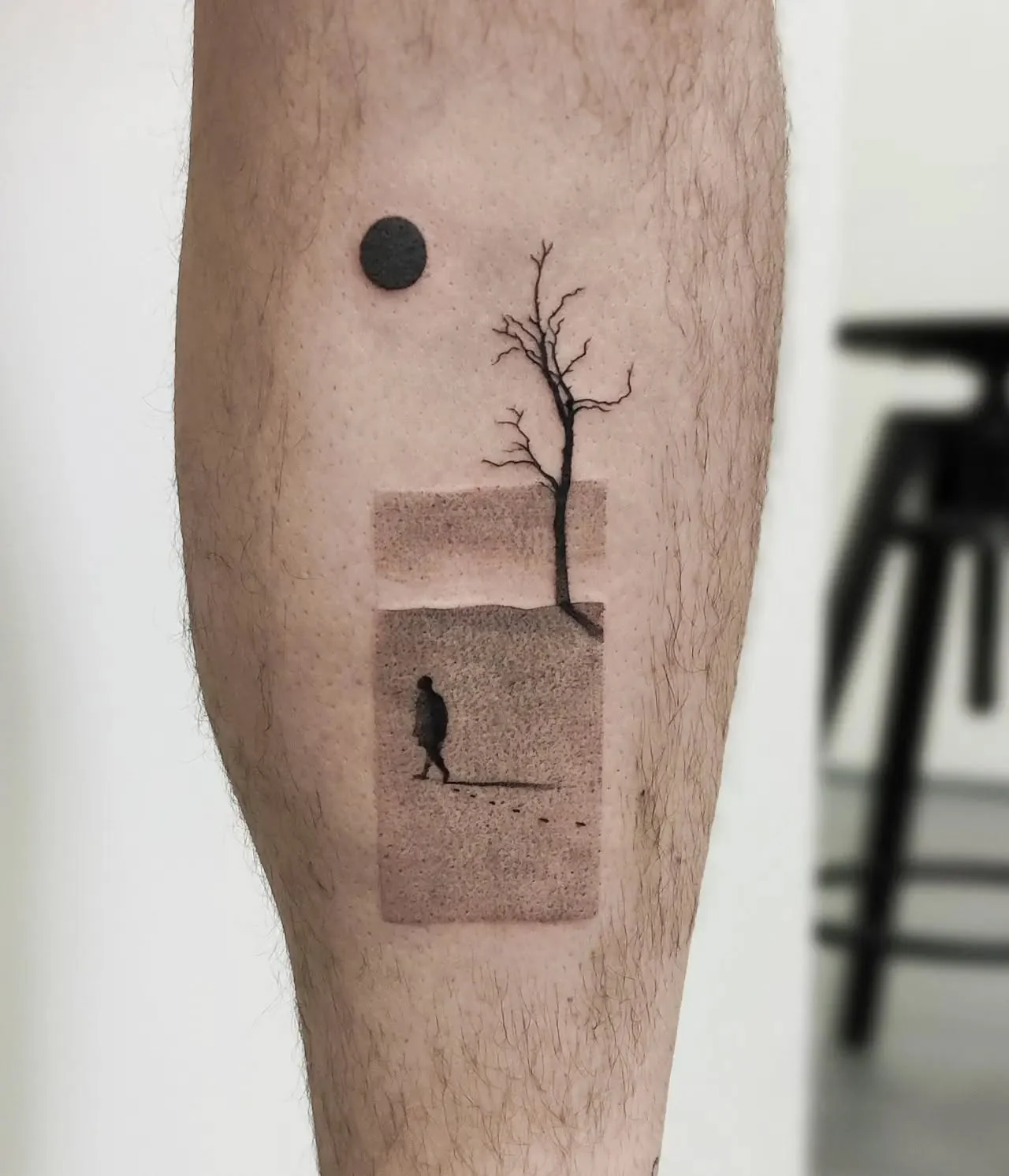 Tatuaż przedstawiający melancholijny krajobraz z bezlistnym drzewem, słońcem lub księżycem oraz sylwetką idącej osoby.