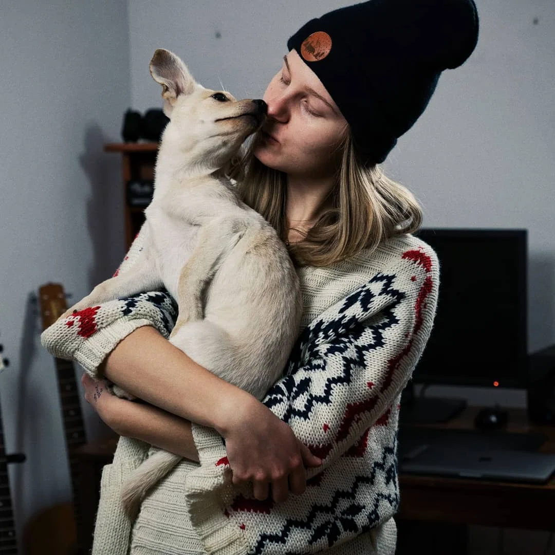 Klara Keczyńska w czarnej czapce i wzorzystym swetrze przytula białego psa, który liże ją po twarzy.