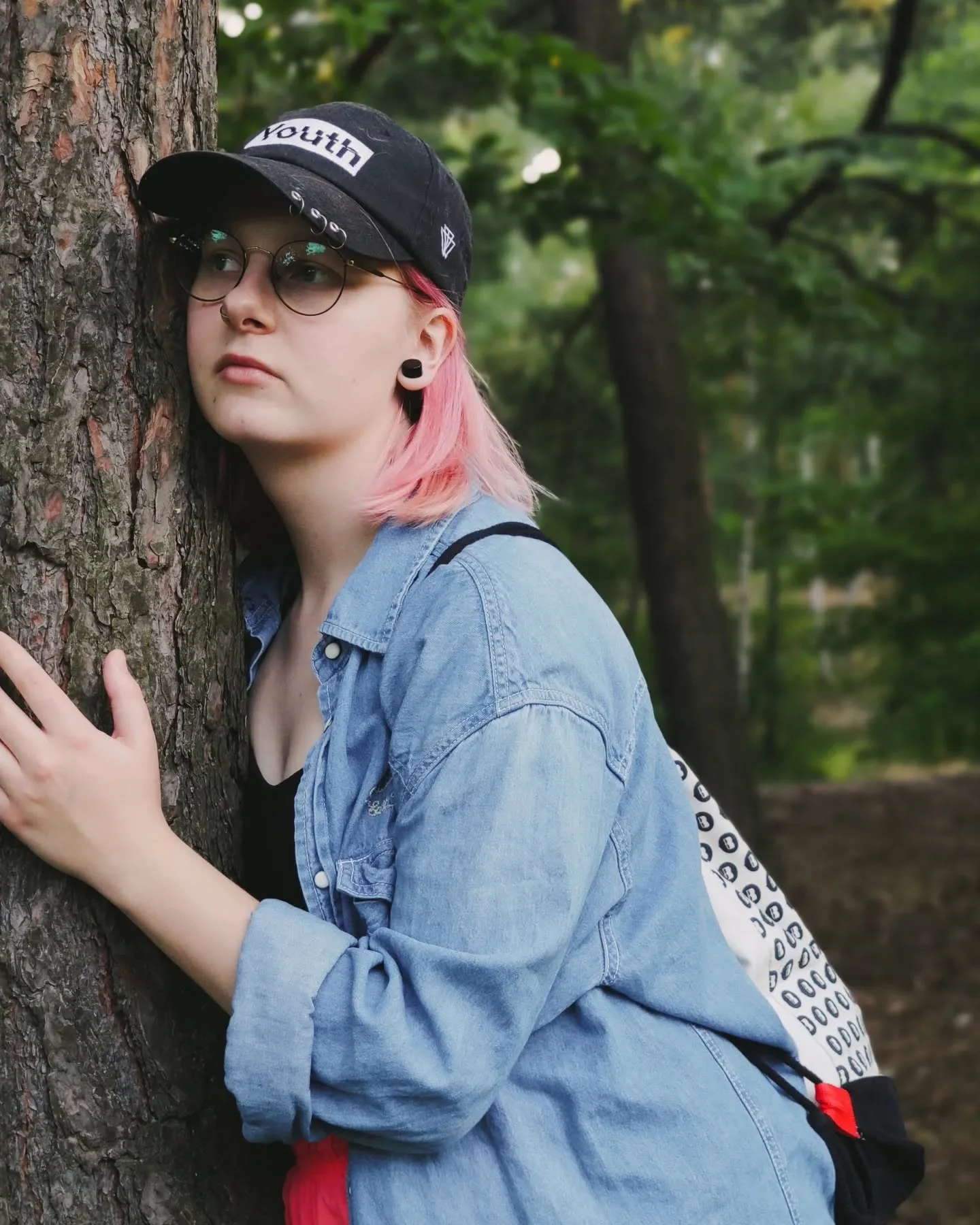 Klara Keczyńska w czapce i okularach opiera się o pień drzewa w lesie.