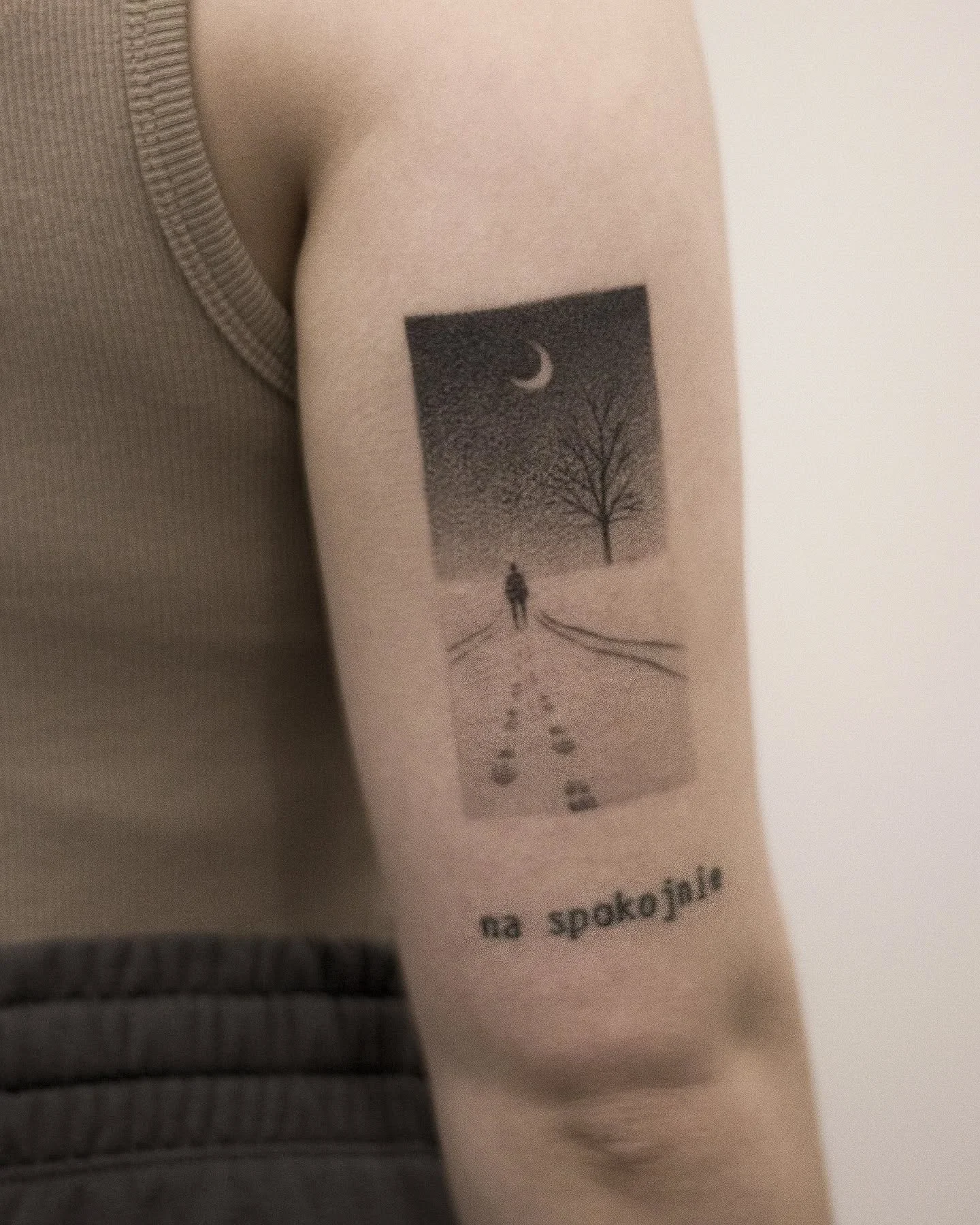 Wygojony tatuaż przedstawiający melancholijny pejzaż z nocnym niebem, półksiężycem, drzewem i postacią idącą ścieżką; poniżej widoczny jest napis „na spokojnie”.