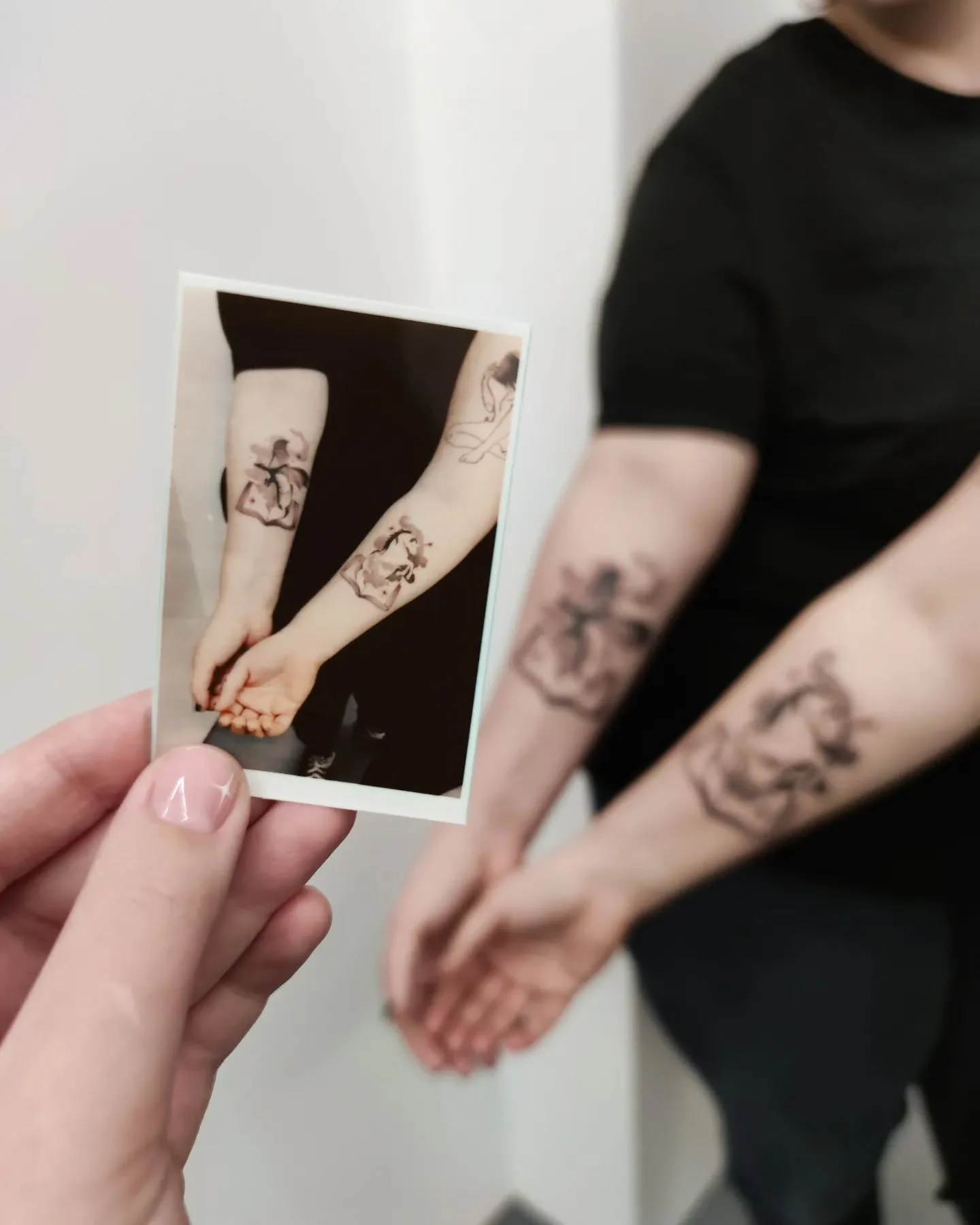 Zdjęcie przedstawia osobę z figuratywnymi tatuażami na przedramionach; na pierwszym planie trzymana jest fotografia tych samych wytatuowanych przedramion.