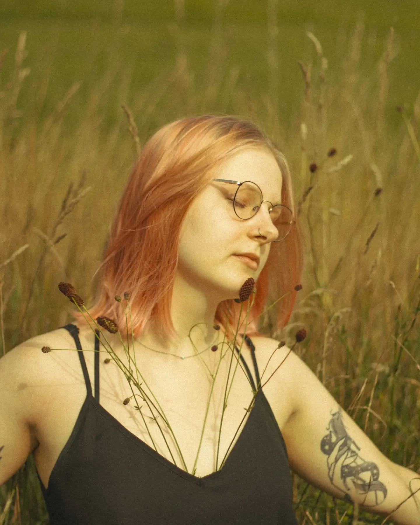 Portret Klary Keczyńskiej w okularach i czarnej sukience, stojącej wśród wysokich traw; na ramionach widoczne są tatuaże., zdjęcie 3 z 4