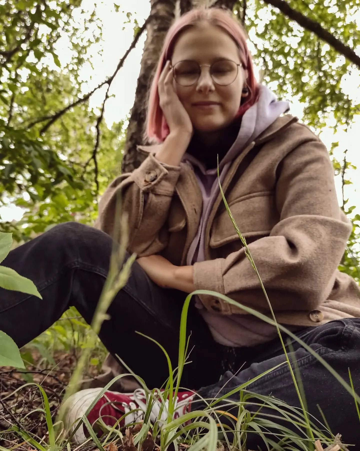Klara Keczyńska siedzi w lesie, oparta o pień drzewa, wśród roślin i leśnej zieleni.