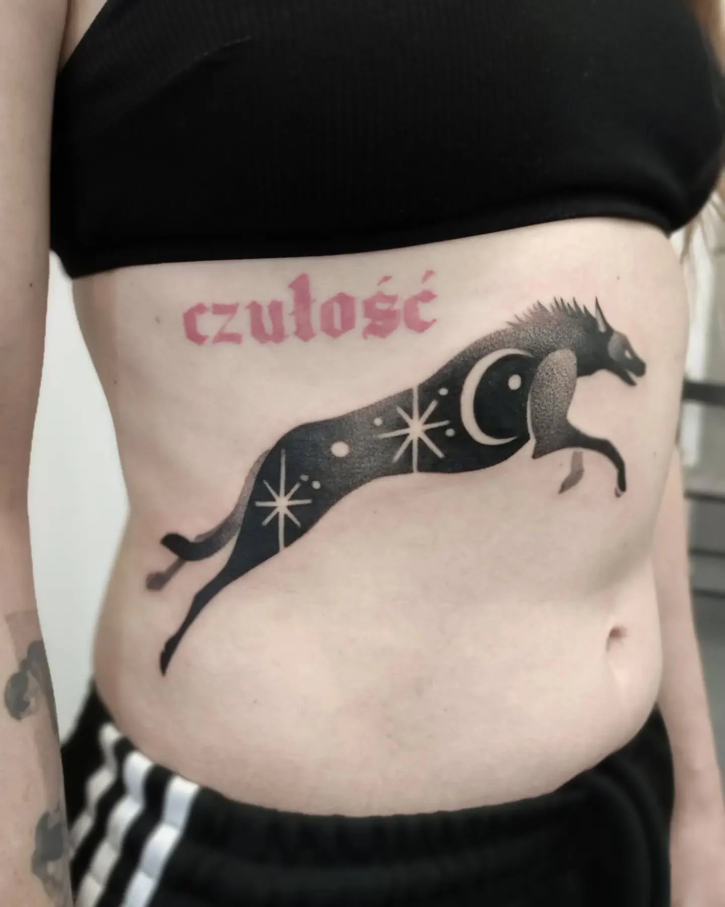 Czarny tatuaż przedstawiający biegnącego wilczura z księżycem i gwiazdami, umieszczony na brzuchu; nad nim widoczny jest napis „czułość”., zdjęcie 3 z 3