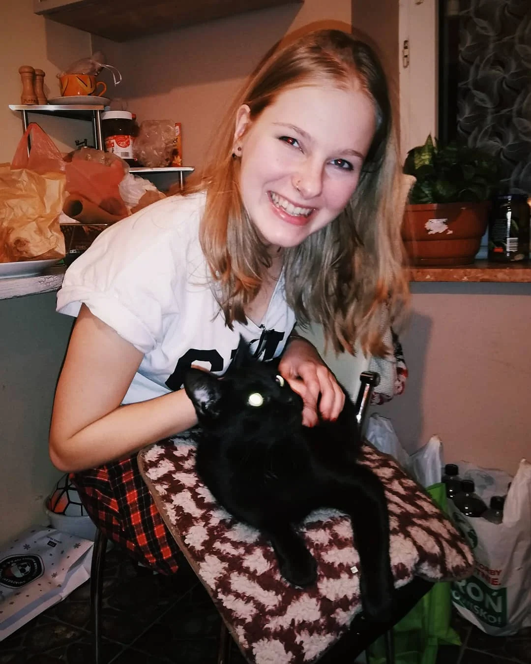 Klara uśmiecha się, trzymając na kolanach czarnego kota w domowym wnętrzu.