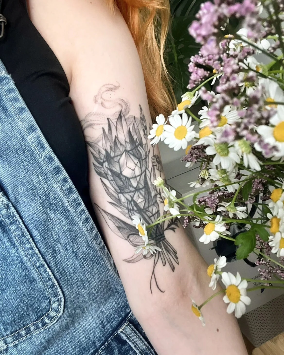 Wygojony cover tatuażu na ramieniu, przedstawiający botaniczną kompozycję z liśćmi, kwiatami i ornamentem; obok widoczny bukiet polnych kwiatów., zdjęcie 1 z 4
