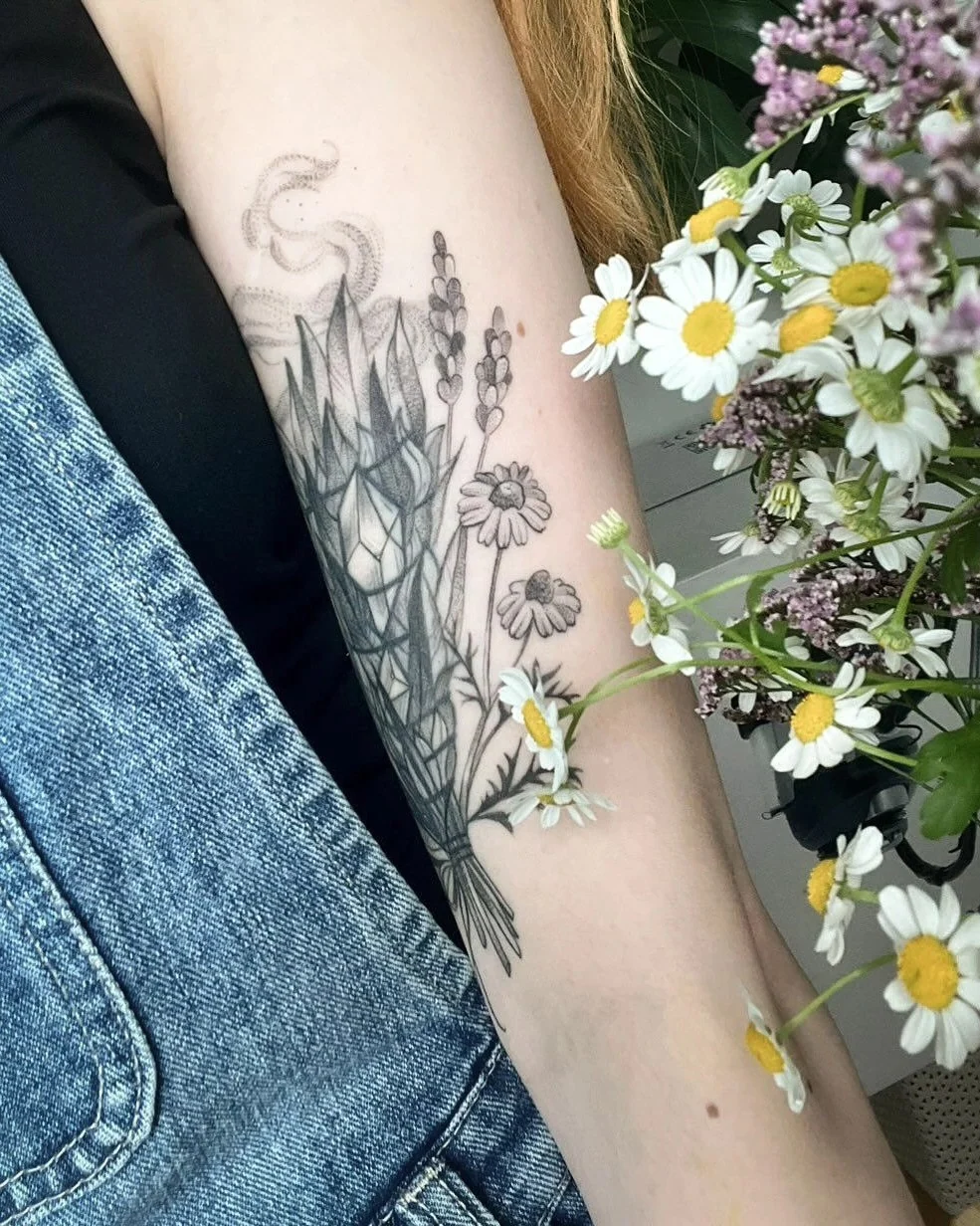 Wygojony cover tatuażu na ramieniu, przedstawiający botaniczną kompozycję z liśćmi, kwiatami i ornamentem; obok widoczny bukiet polnych kwiatów., zdjęcie 3 z 4