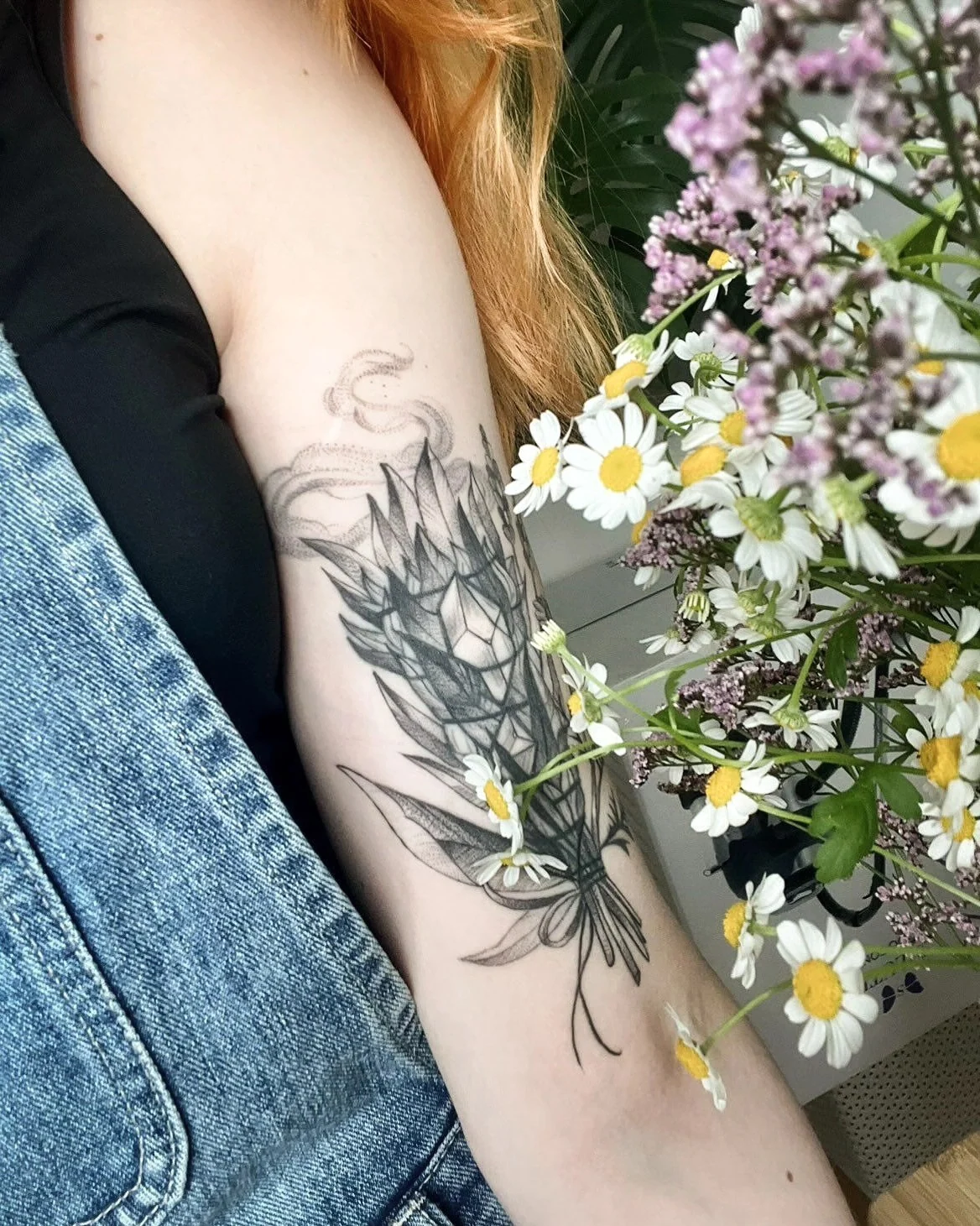 Wygojony cover tatuażu na ramieniu, przedstawiający botaniczną kompozycję z liśćmi, kwiatami i ornamentem; obok widoczny bukiet polnych kwiatów., zdjęcie 2 z 4