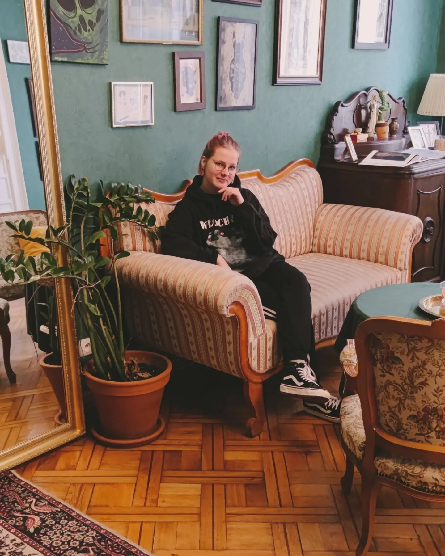 Klara Keczyńska siedzi na wzorzystej kanapie w ozdobnym wnętrzu z roślinami i obrazami na ścianach.