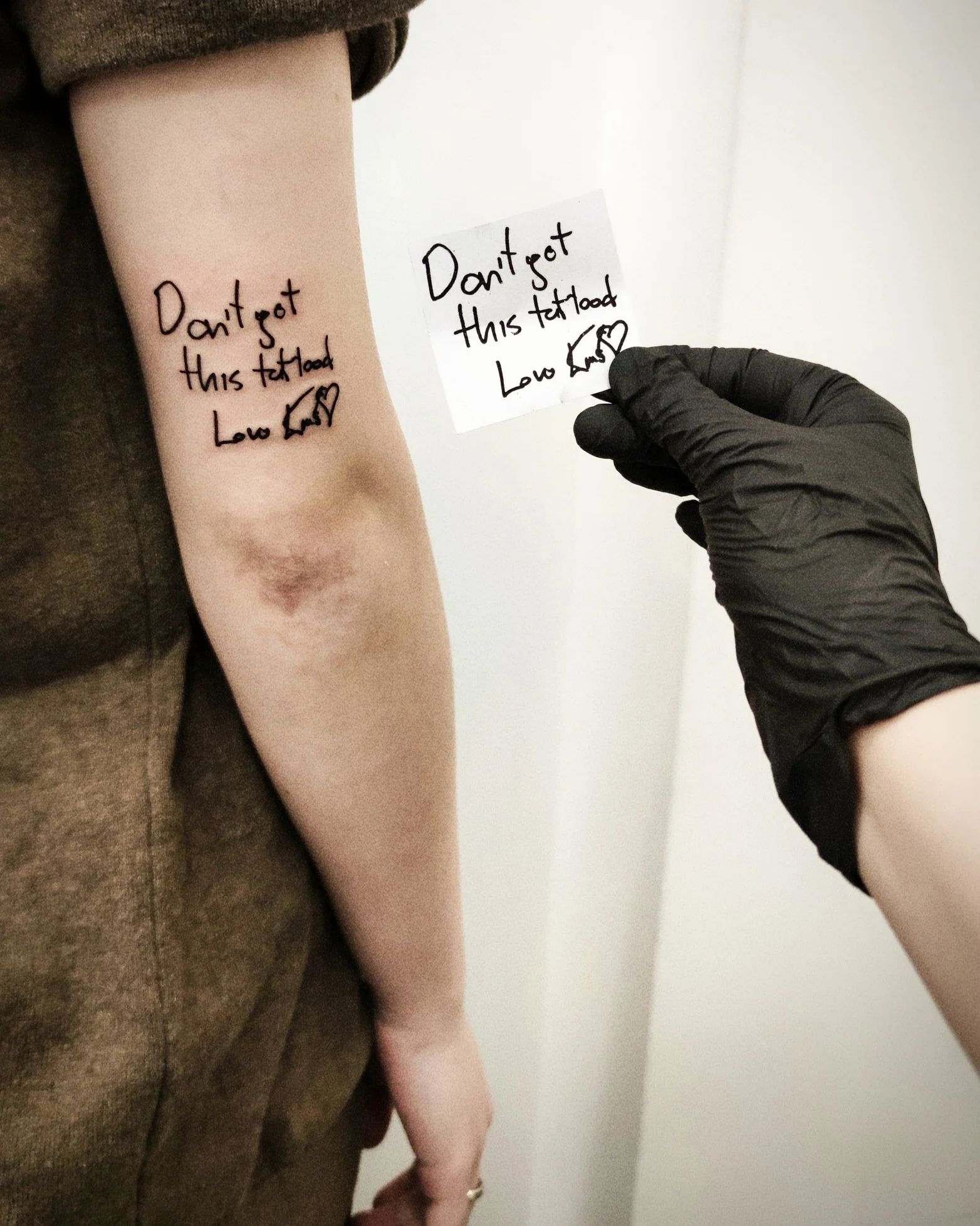 Zbliżenie czarnego tatuażu z odręcznym napisem „Don’t got this tattooed”, małym symbolem serca i dodatkową ilustracją; na pierwszym planie widać ramię oraz kartkę z projektem napisu., zdjęcie 1 z 2