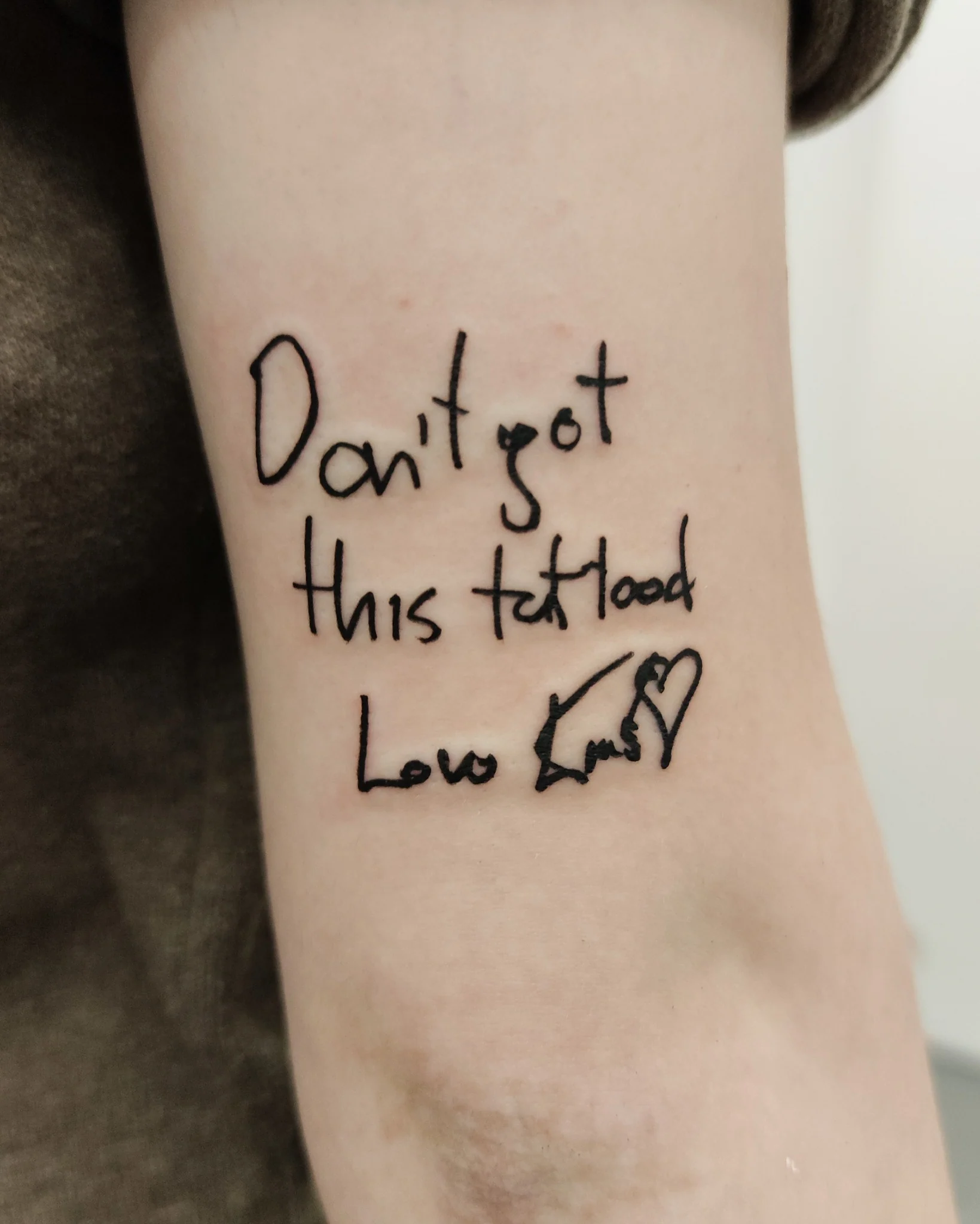 Zbliżenie czarnego tatuażu z odręcznym napisem „Don’t got this tattooed”, małym symbolem serca i dodatkową ilustracją; na pierwszym planie widać ramię oraz kartkę z projektem napisu., zdjęcie 2 z 2