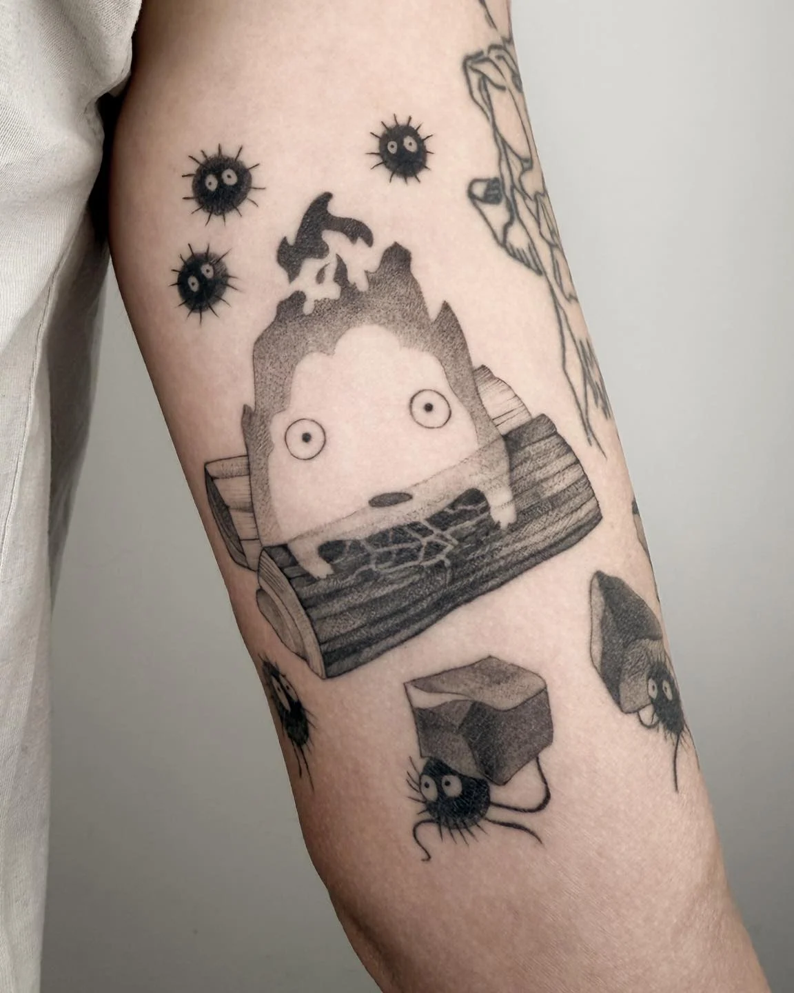 Wygojony tatuaż na ramieniu przedstawiający Calcifera oraz małe czarne kurzyki inspirowane filmami Studia Ghibli., zdjęcie 1 z 4
