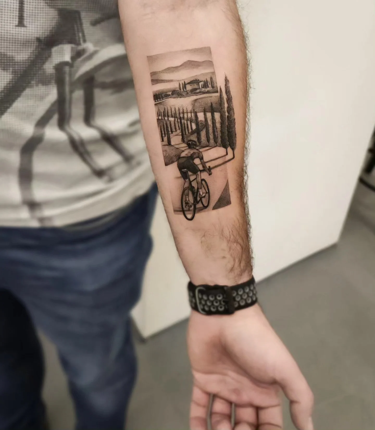 Tatuaż przedstawiający rowerzystę jadącego przez krajobraz z cyprysami, wzgórzami i drogą, wykonany na przedramieniu., zdjęcie 2 z 3