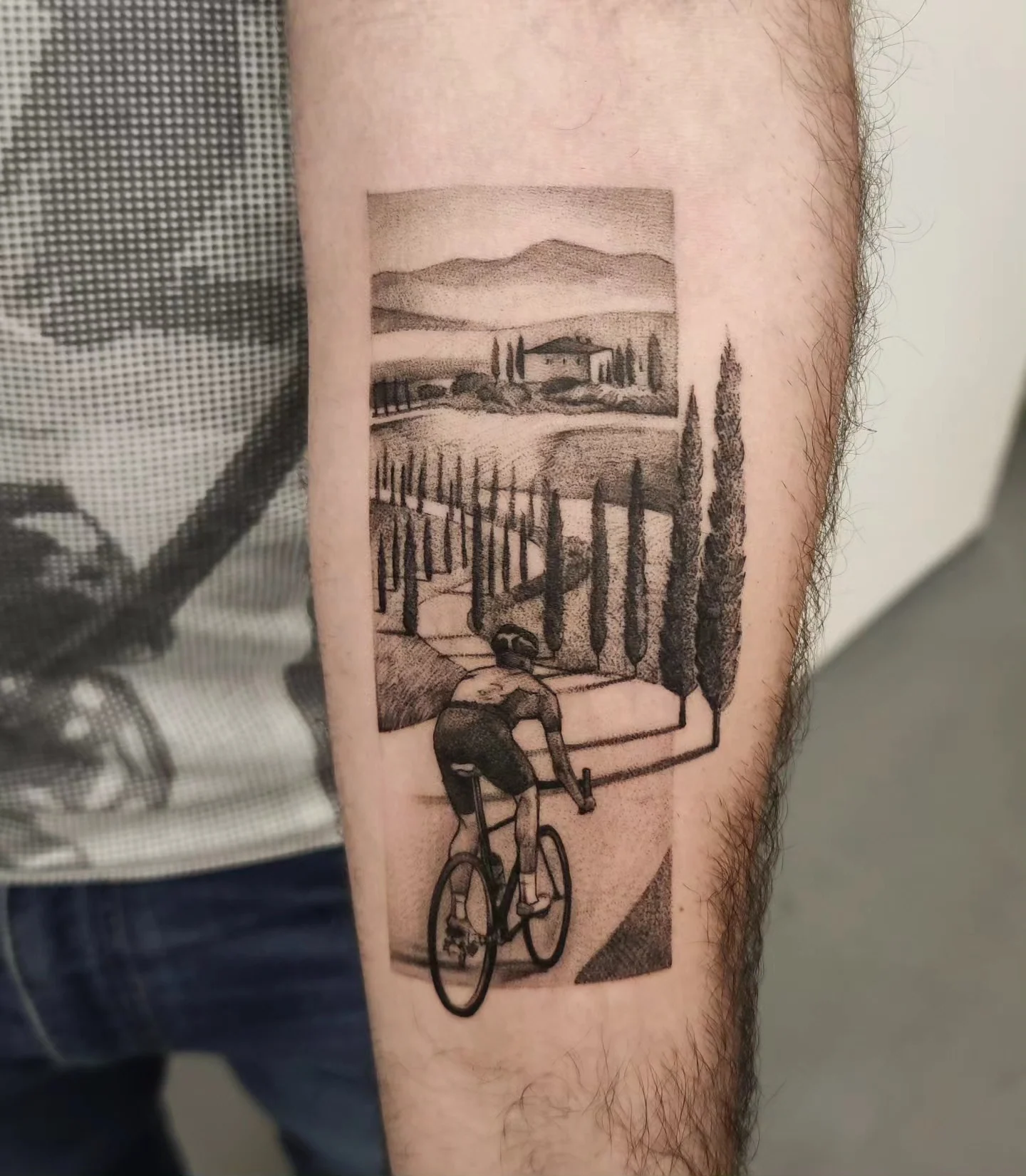 Tatuaż przedstawiający rowerzystę jadącego przez krajobraz z cyprysami, wzgórzami i drogą, wykonany na przedramieniu., zdjęcie 1 z 3