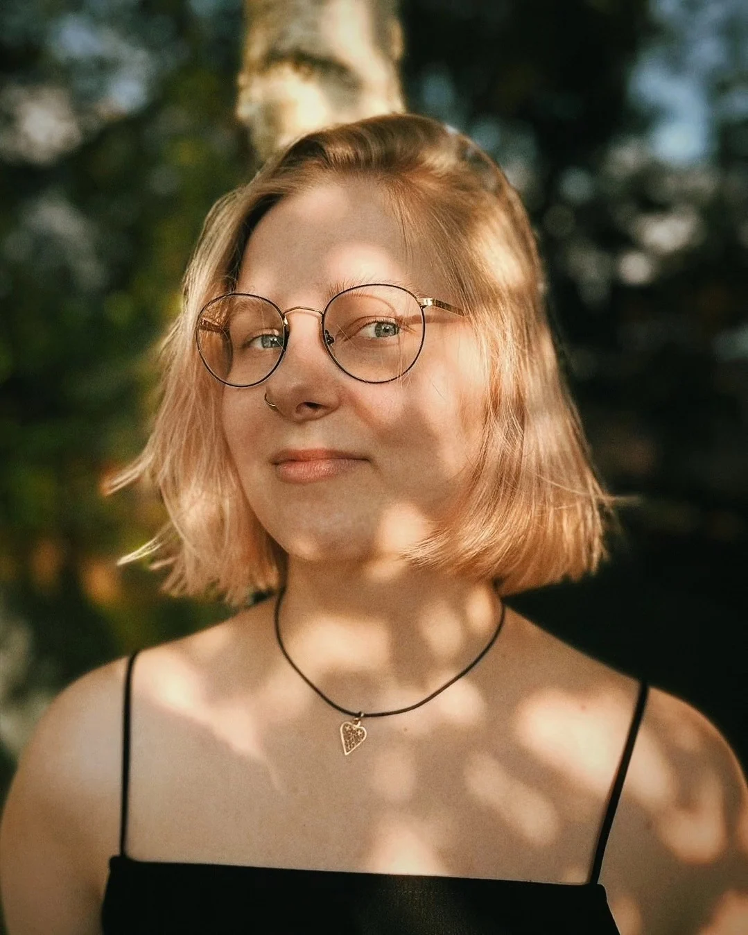 Portret Klary Keczyńskiej w okularach i czarnej sukience, sfotografowanej wśród drzew w naturalnym świetle.